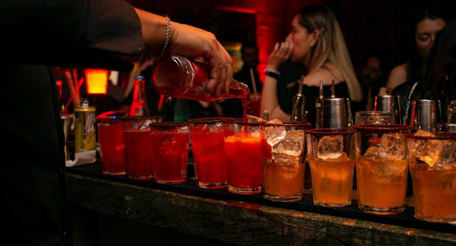 Un bar tender sirviendo de noche una hielera de vasos con cócteles de color rojo vibrante y naranja con hielo, bajo una iluminación cálida en un bar en Cholula Puebla.